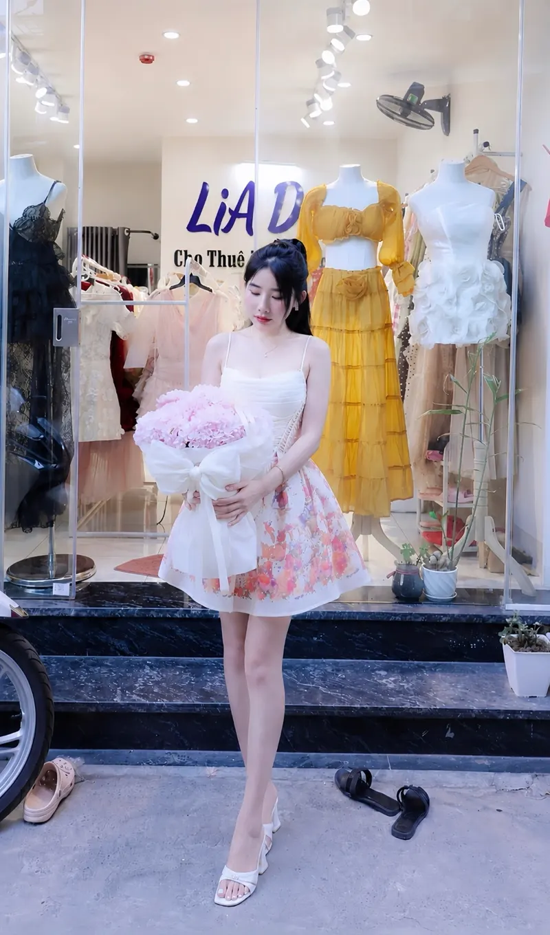Nàng thơ LiA Dress mặc đầm hai dây hoạ tiết hoa pastel, tay ôm bó hoa cẩm tú cầu hồng trước showroom.