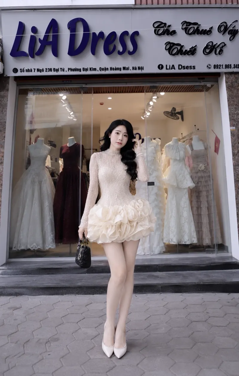 Người mẫu diện đầm cổ cao ren kem với chân váy bồng hoa hồng organza, đứng trước biển hiệu LiA Dress.